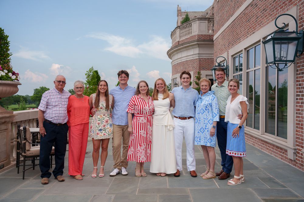 A large family group poses together smiling on an outdoor stone patio of an elegant brick mansion.