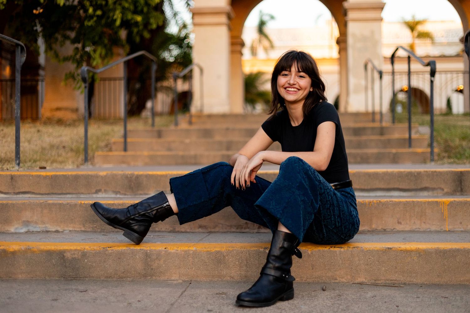 A high school senior in black top and jeans sitting casually on outdoor stone steps with arches in background at sunset.