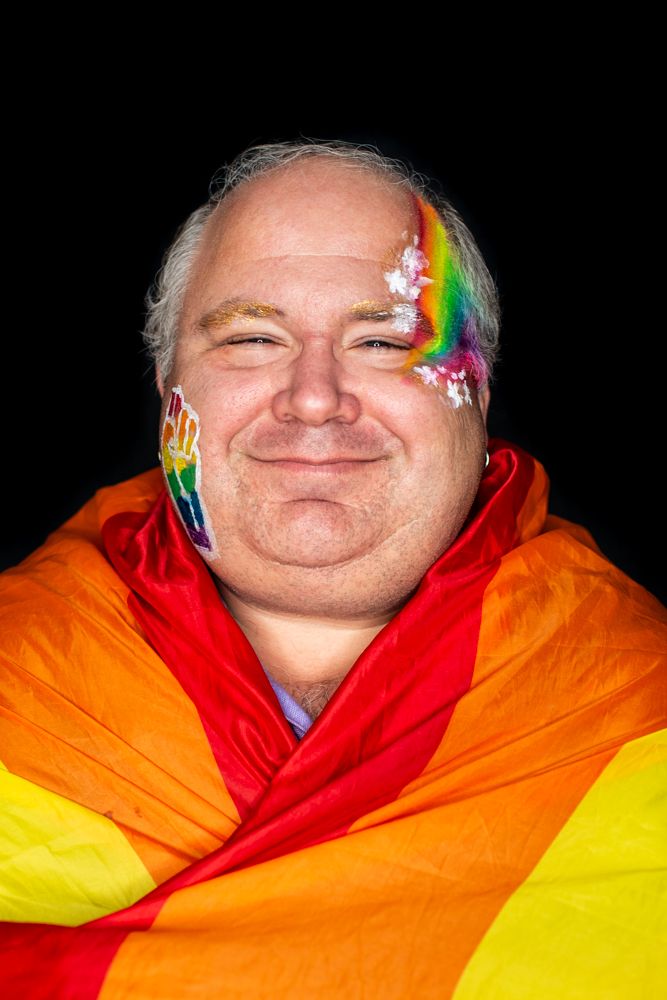 A person wrapped in a rainbow pride flag with rainbow face paint smiles against a dark background.