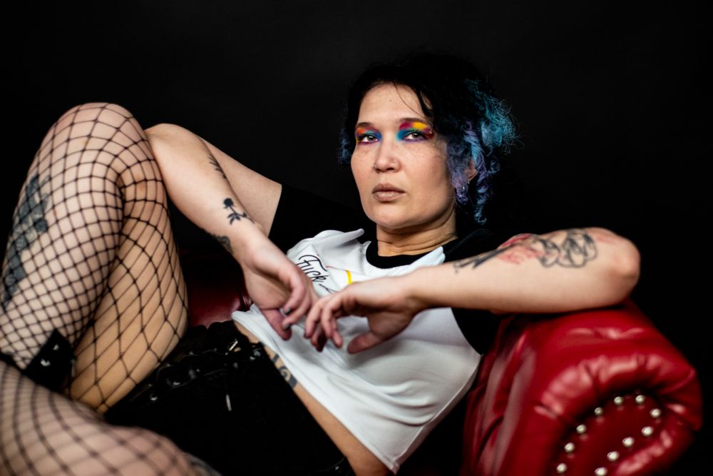 Alternative style portrait with subject wearing fishnet stockings lounging on red leather couch with dramatic lighting.