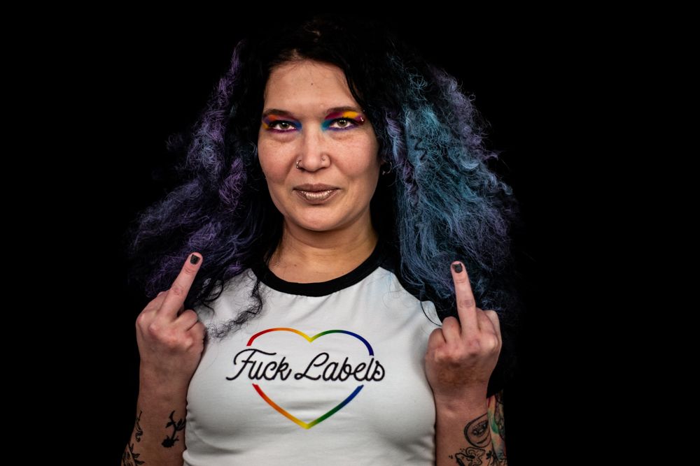 Person wearing a heart rainbow t-shirt against black background giving double middle finger gesture with colorful hair.