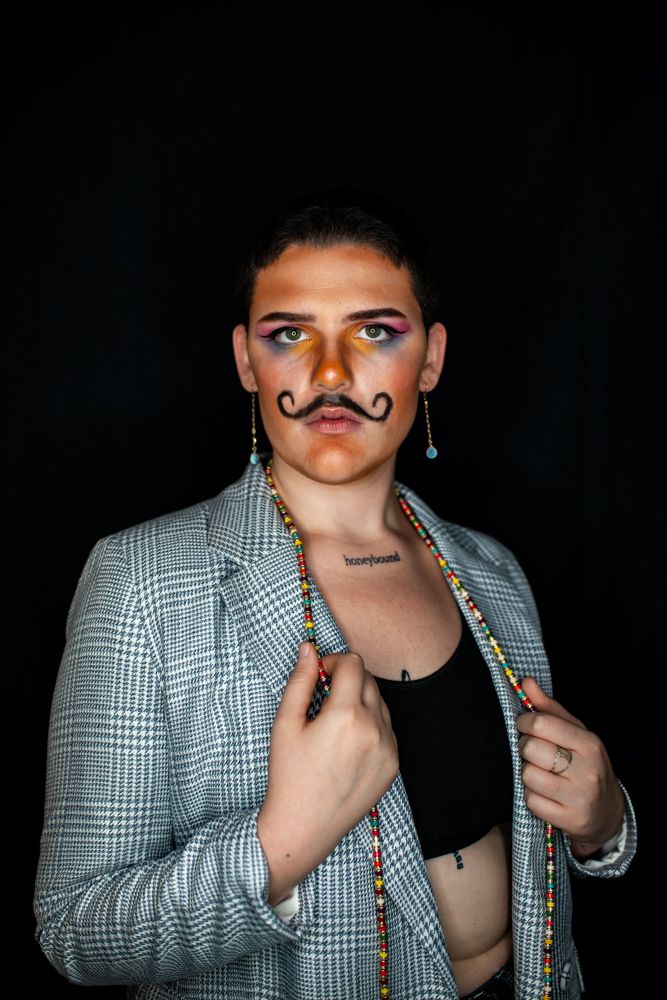 A figure wearing a grey plaid blazer strikes different poses against a black background with drawn-on mustache and props.