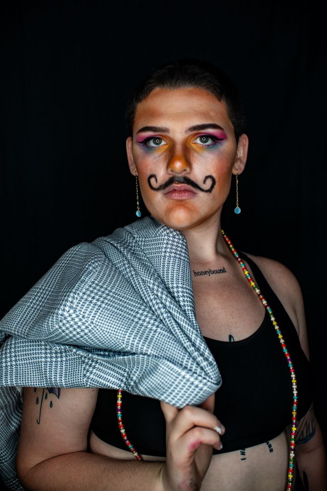A creative photoshoot against black background features artful makeup and a checkered pattern shirt.
