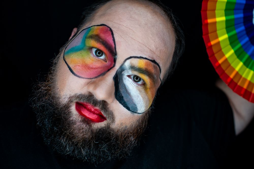 Creative pride-themed makeup look with rainbow colors, black and white contrast, red lipstick, and beard against dark background.