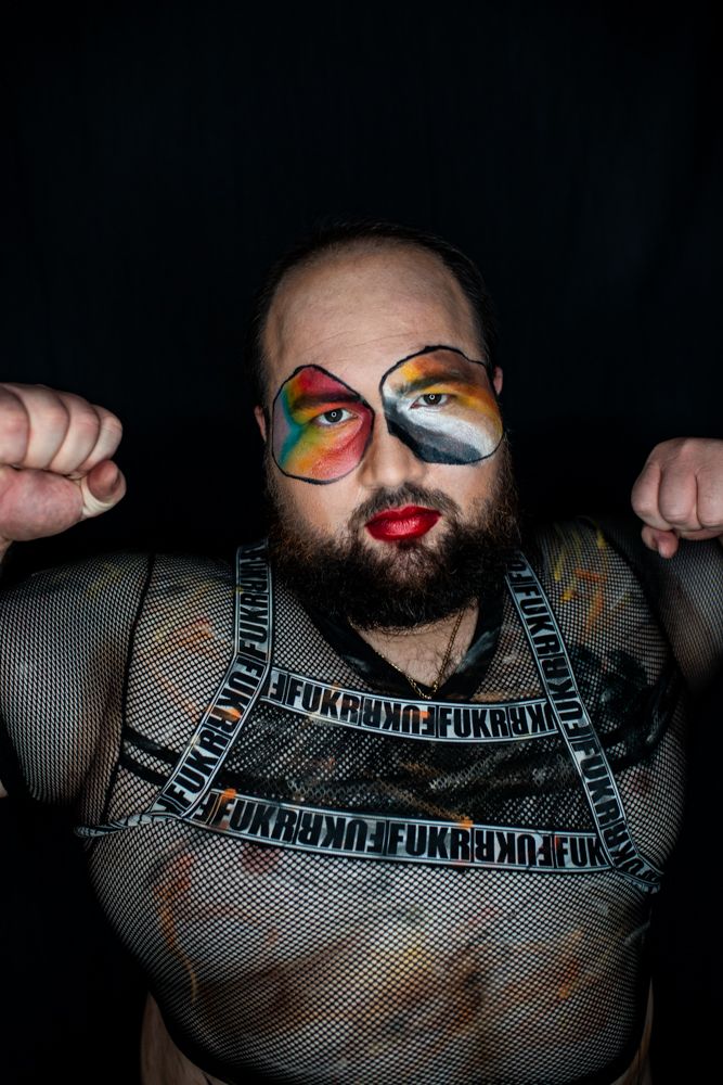 Bearded person wearing colorful face paint and mesh harness poses with raised fists against dark background.