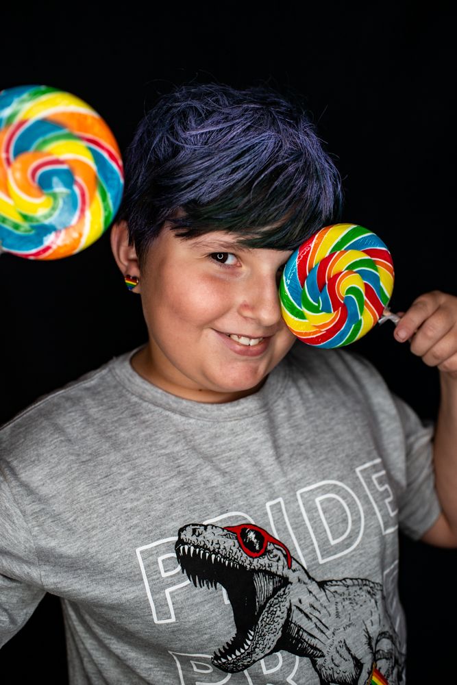 A person in a grey dinosaur t-shirt holds two colorful rainbow lollipops against a black background.