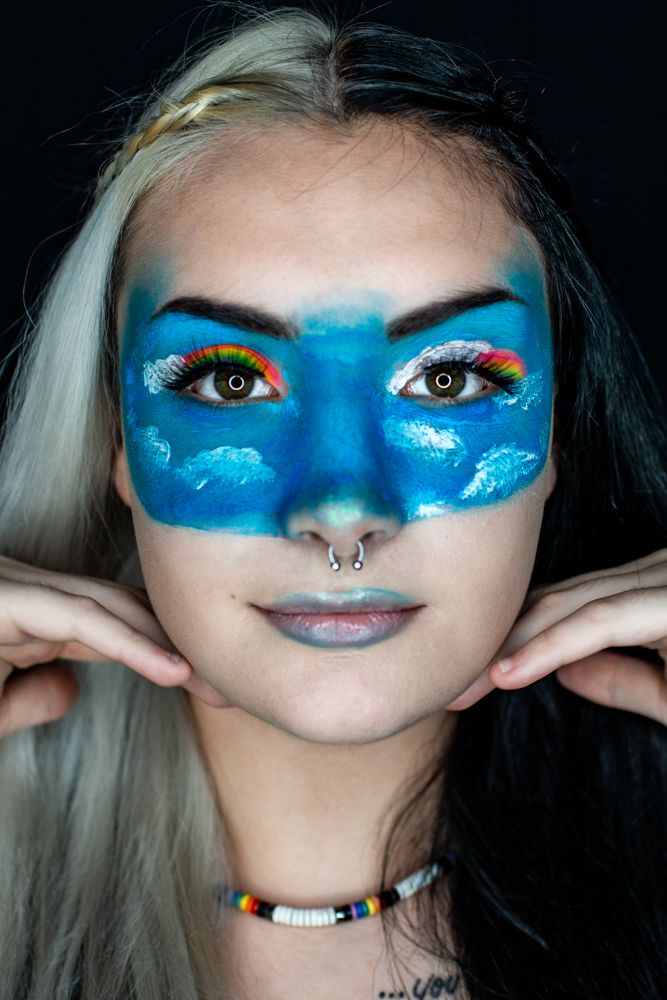 Creative sky-inspired makeup art features bright blue face paint with white cloud details and vibrant orange eyeshadow.