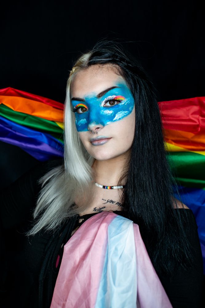 Creative portrait with blue face paint and two-toned hair against rainbow pride flag background.