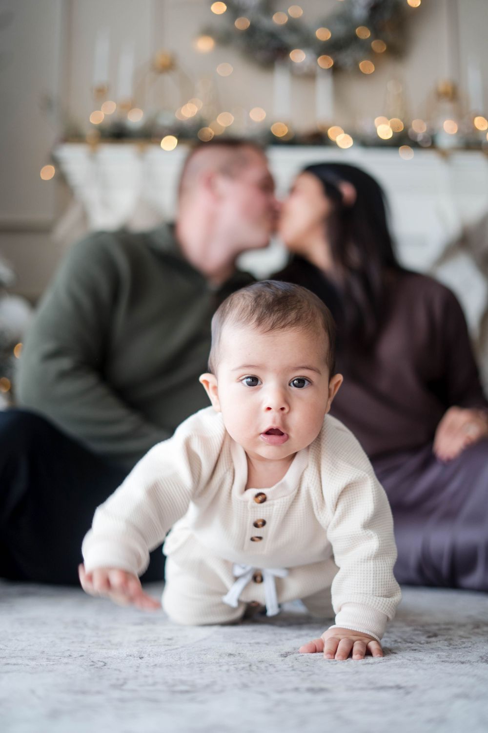 Curious baby in white onesie crawls on the floor while parents share a kiss during Christmas photos.