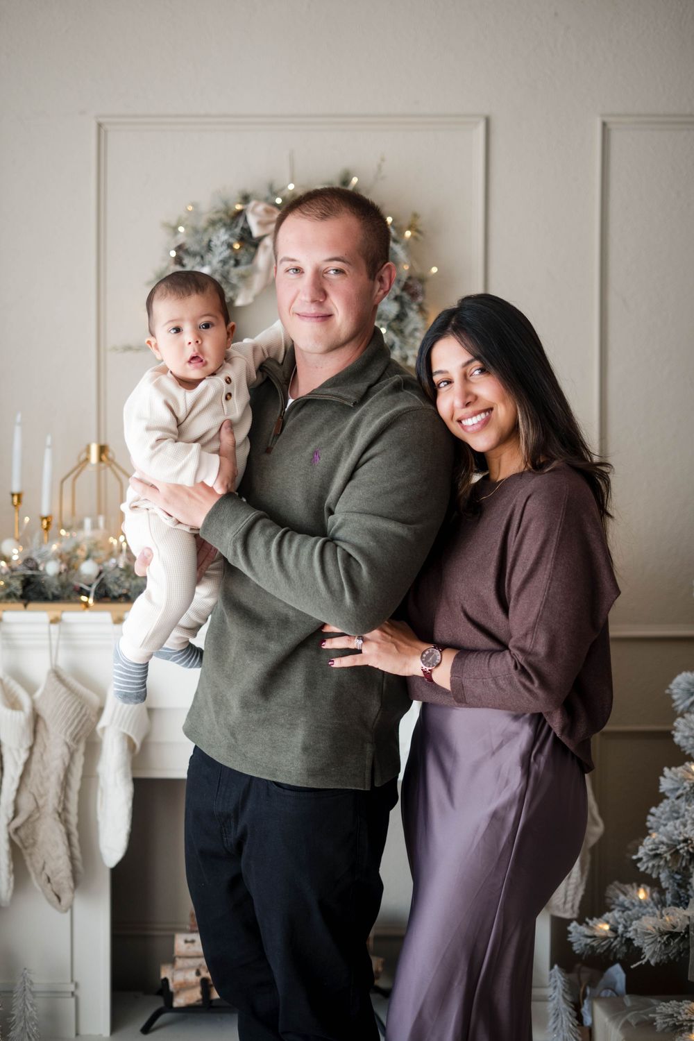 Young family with a baby in a cream outfit poses warmly in front of a Christmas fireplace mantel.