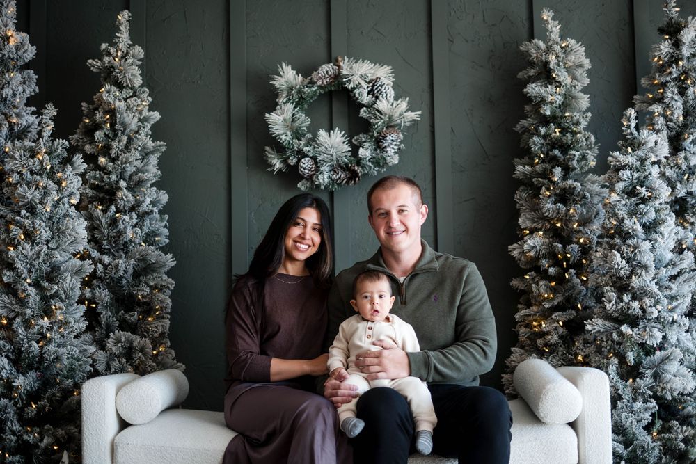 Couple in brown and green outfits sits on a white sofa holding their baby between snowy Christmas trees.