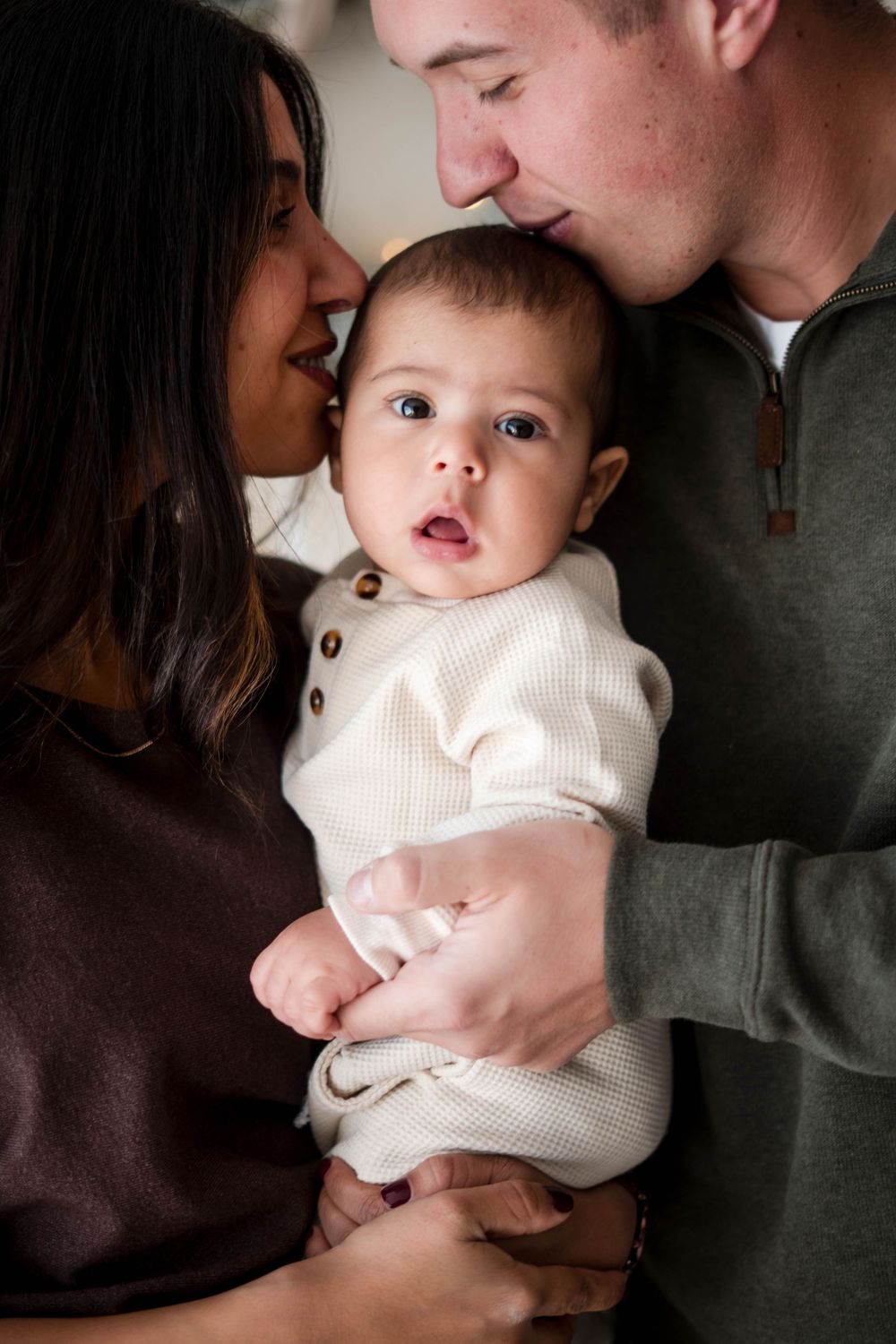 New parents in brown and green outfits tenderly kiss their wide-eyed baby during a holiday photo session.