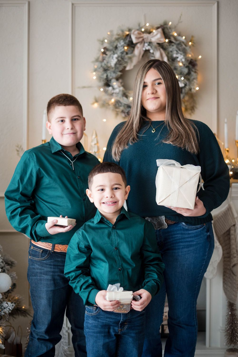 Two boys in green shirts and a teenage girl holding Christmas gifts pose in front of a holiday wreath.
