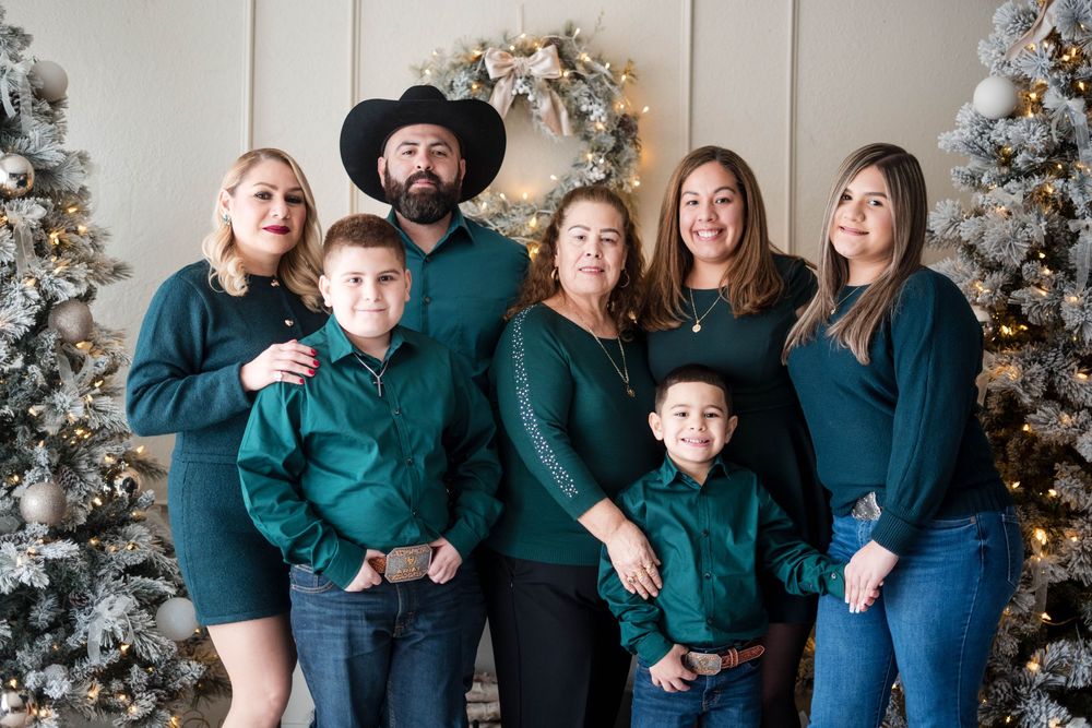 Family of seven in matching green outfits posing together in front of decorated Christmas trees for a holiday portrait.
