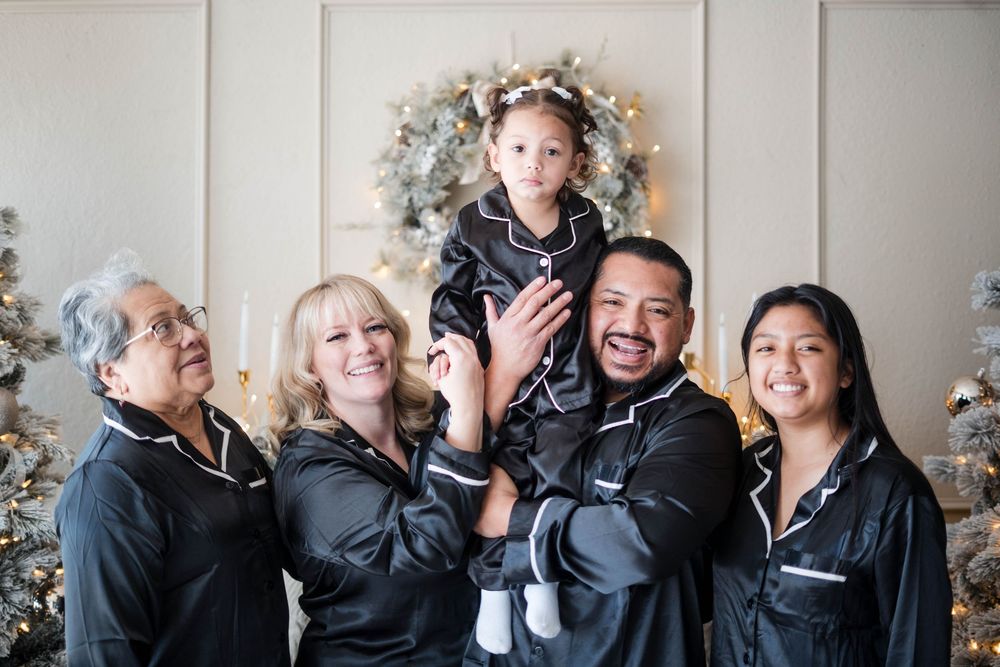 Multigenerational family in black pajamas laughing while lifting a young girl during Christmas portraits.