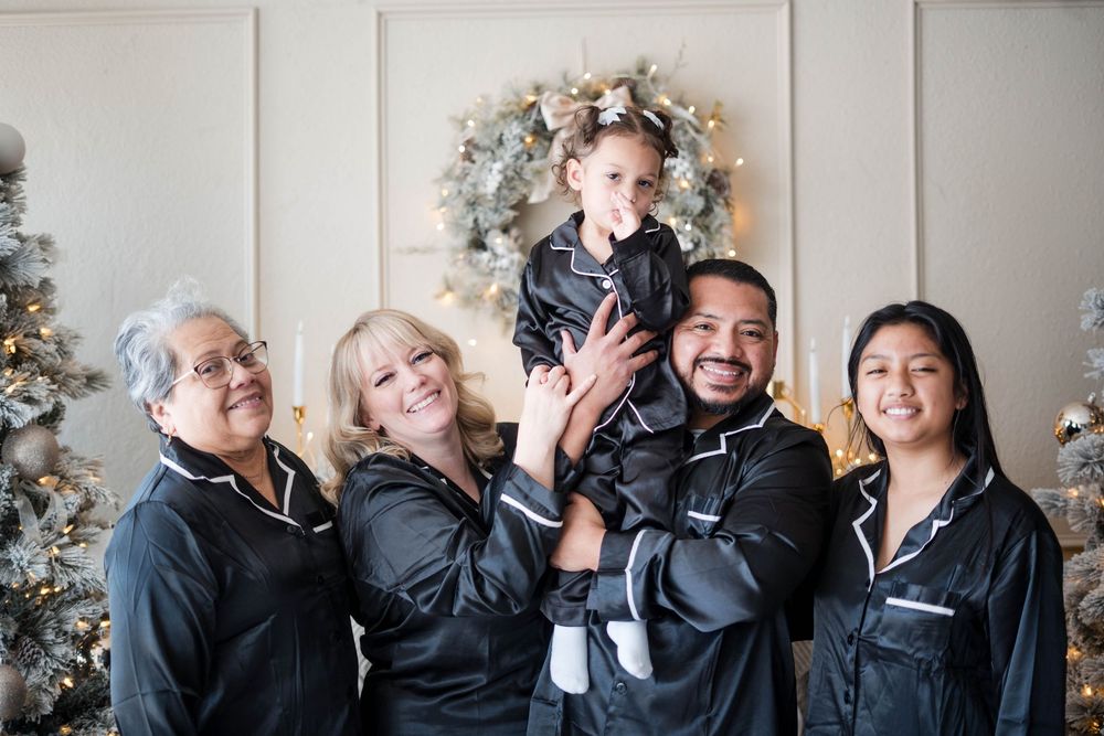 Family of five in matching black satin pajamas posing joyfully with a toddler in front of Christmas trees.