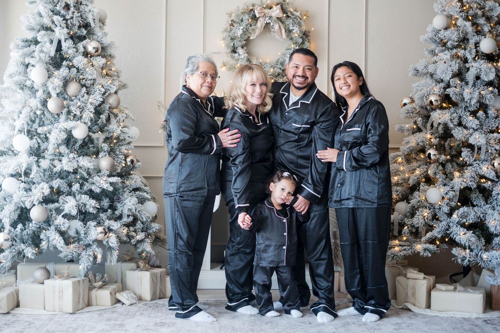 Family of five wearing matching black satin pajamas standing together between decorated Christmas trees.