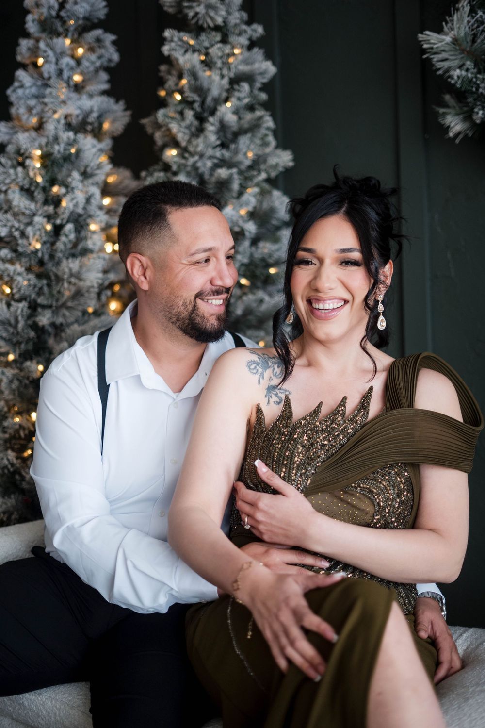 Smiling couple sitting together in front of flocked Christmas trees in elegant holiday attire.