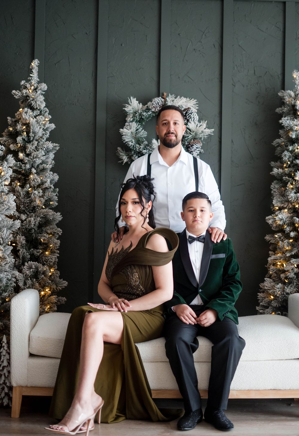 Elegant family of three posing on a white sofa between flocked Christmas trees indoors.