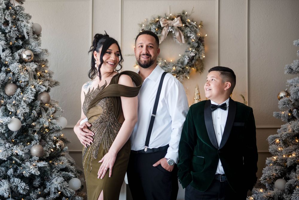 Pregnant woman, man with suspenders, and boy in tuxedo posing by Christmas trees indoors.