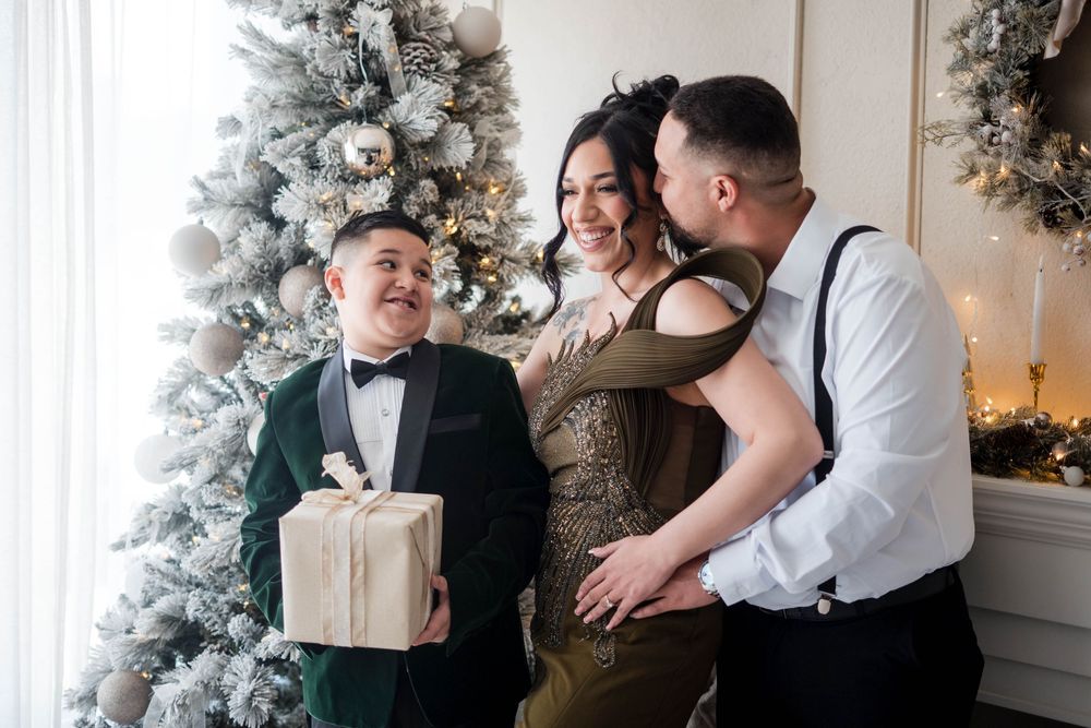 Family of three laughing and sharing a gift beside a decorated Christmas tree indoors.