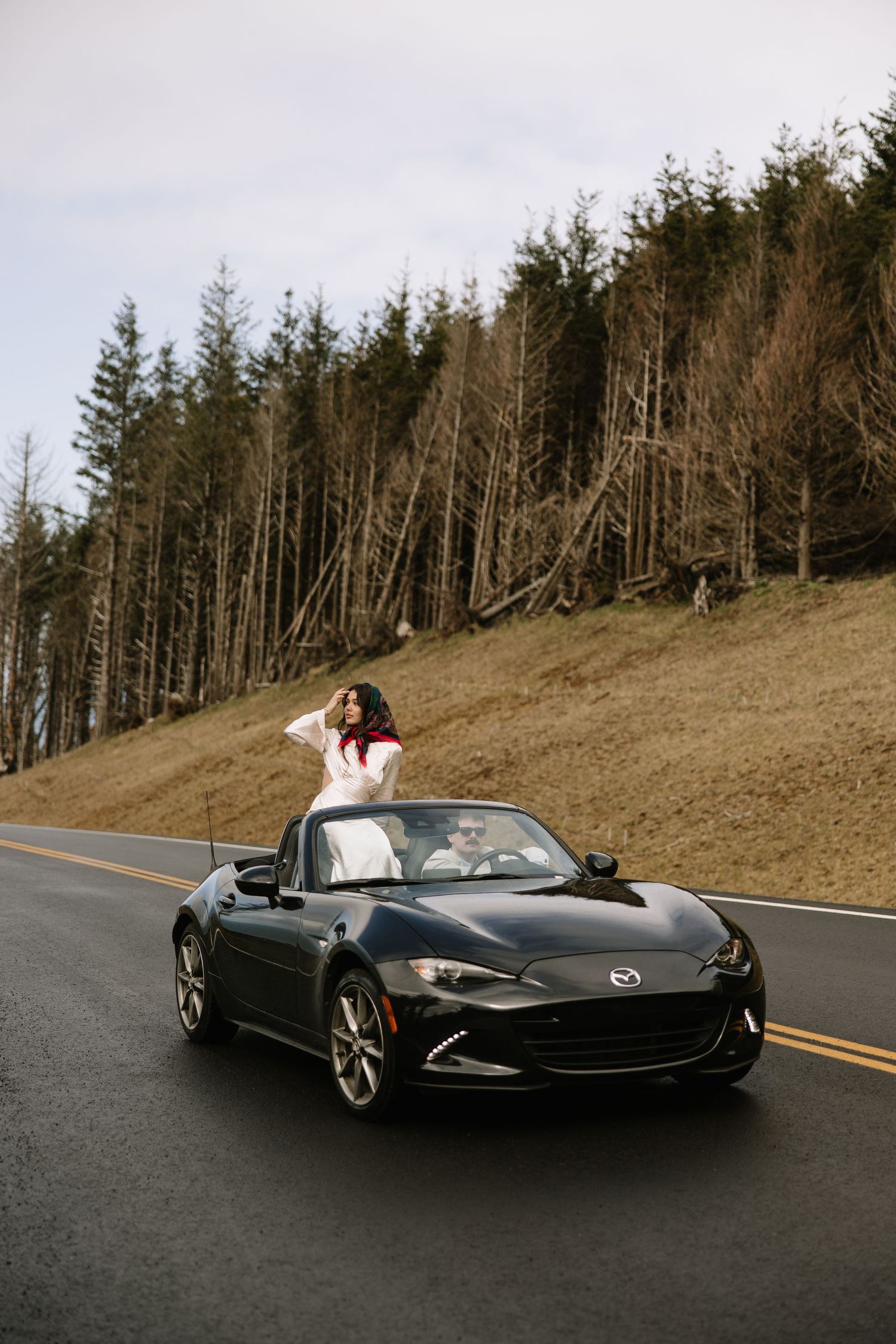 A black Mazda Miata convertible on a curving mountain road with evergreen trees in the background.