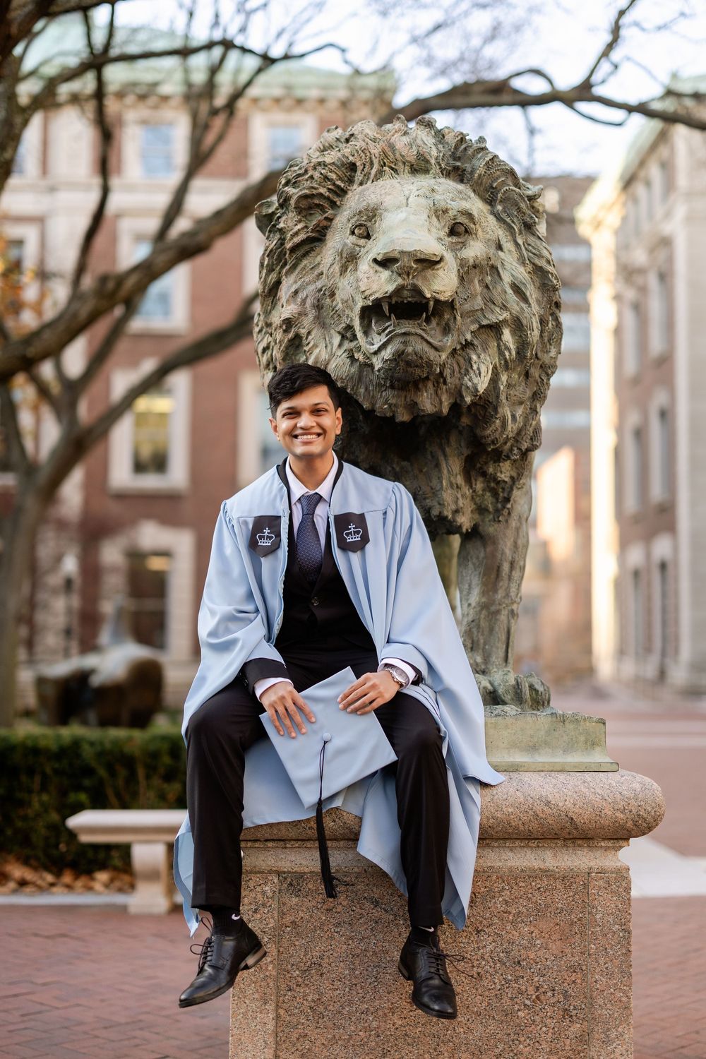 A graduate in a light blue gown sits on a stone ledge near a bronze lion statue on a college campus.
