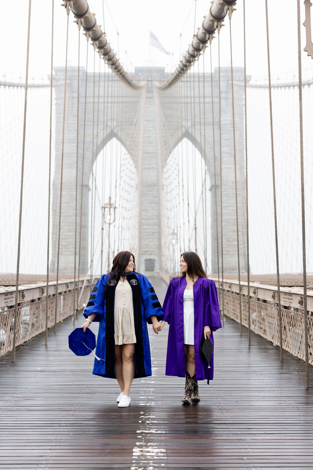 Two people in matching blue and purple jackets walk away on the Brooklyn Bridge on a foggy day.