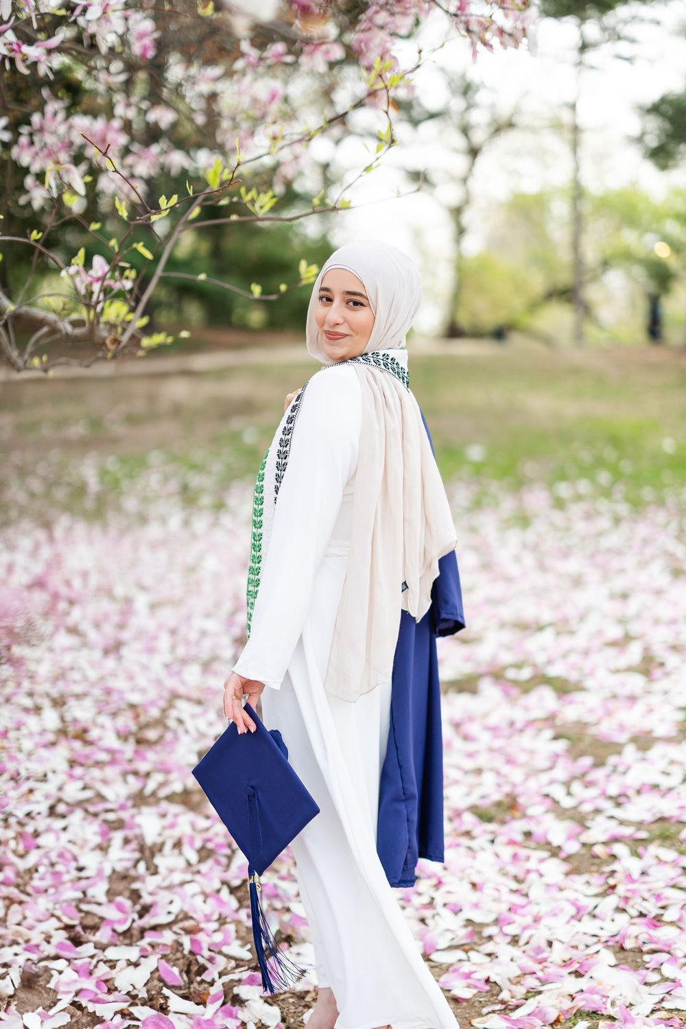 A person in white hijab and navy blue dress poses gracefully among pink cherry blossom petals in a spring garden.