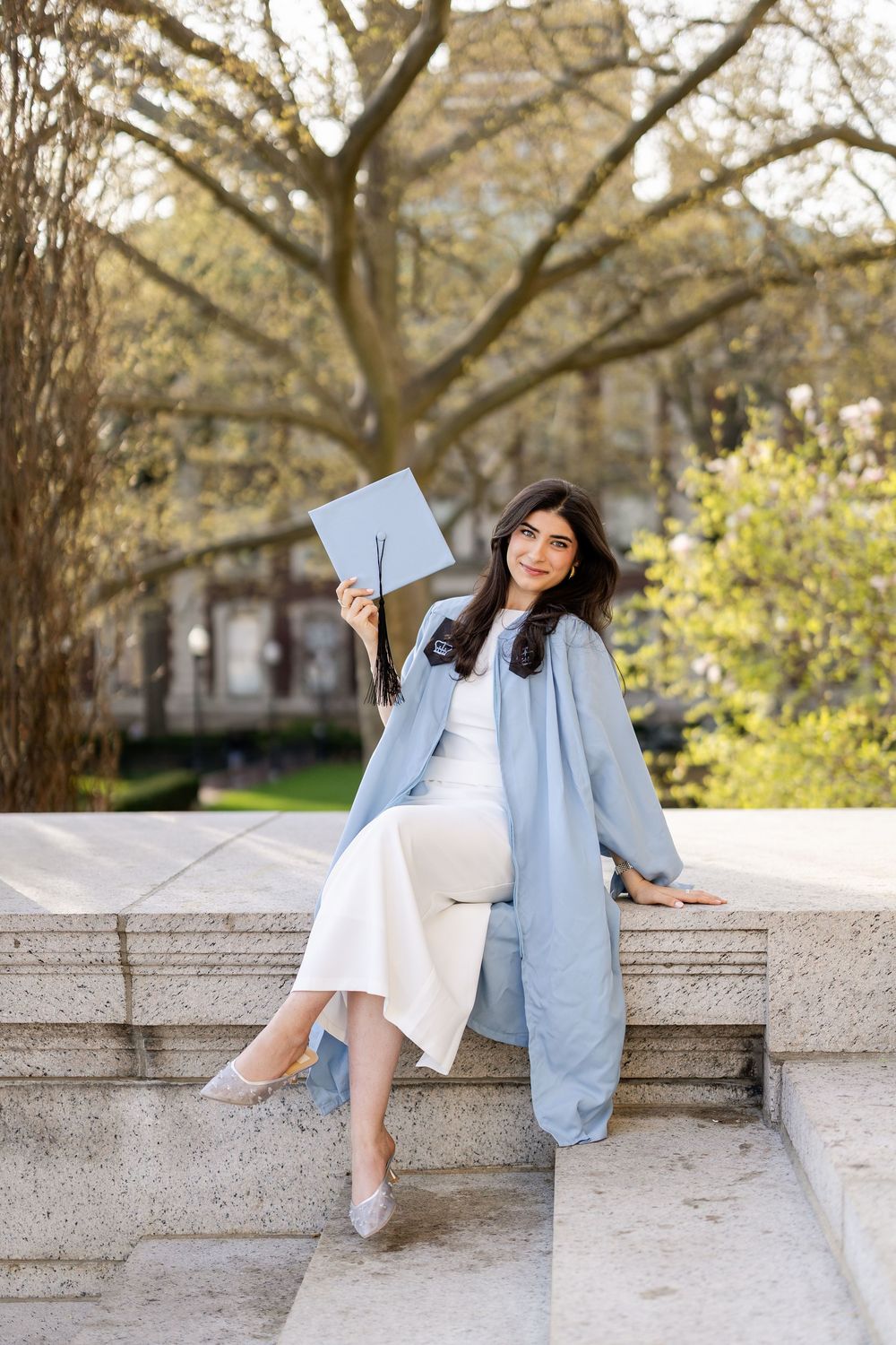 Graduate in light blue coat sits on stone steps holding diploma while posing for graduation photos on campus.