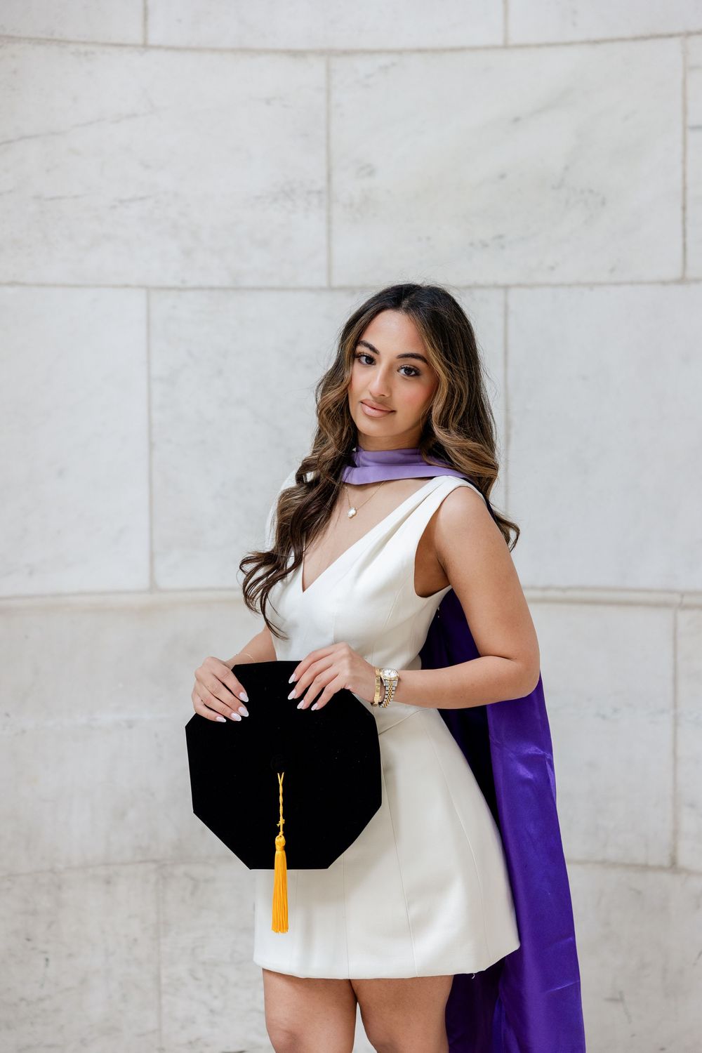 Graduate in white dress and purple graduation stole poses against marble wall in outdoor photoshoot series.