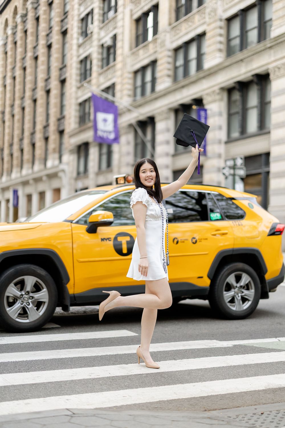 Graduate celebrates in white dress jumping on crosswalk with yellow NYC taxi in background.