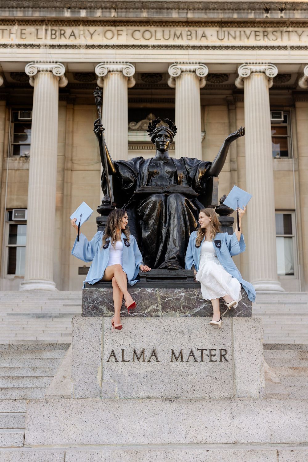 Students in graduation attire pose playfully on the steps of Columbia University's iconic library building.