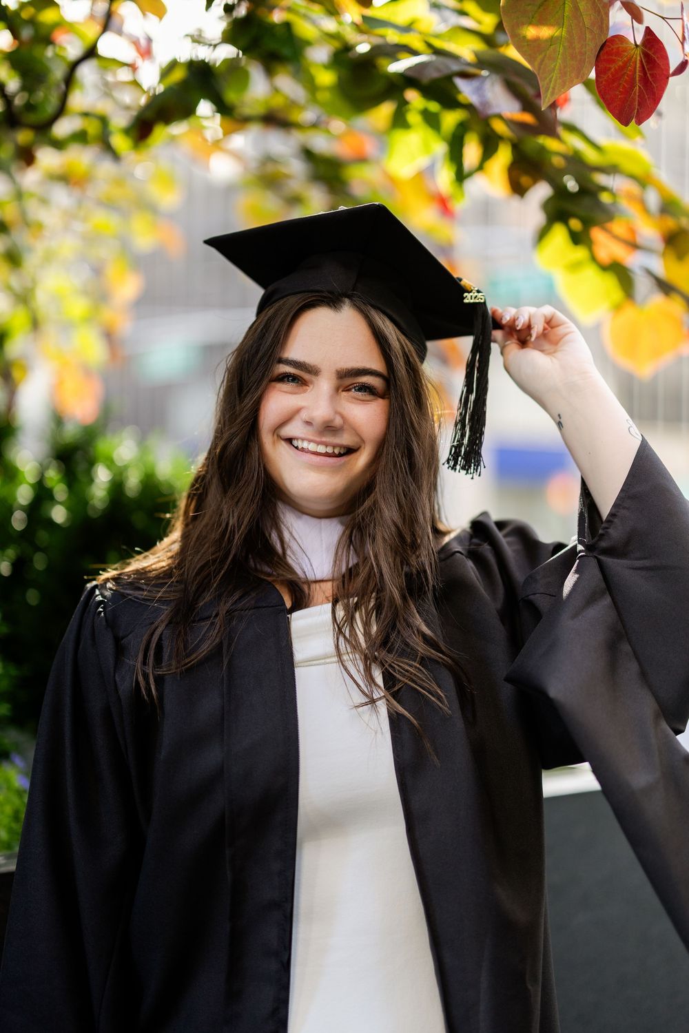 Graduate in cap and gown smiling outdoors under colorful autumn foliage during graduation photo session.