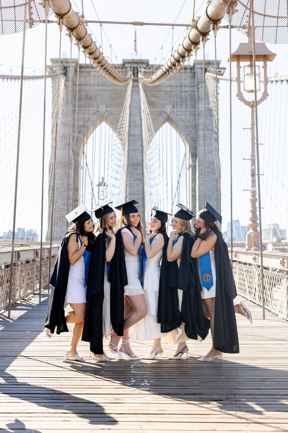 A group of graduates in black caps and gowns pose together on the Brooklyn Bridge walkway in New York City.
