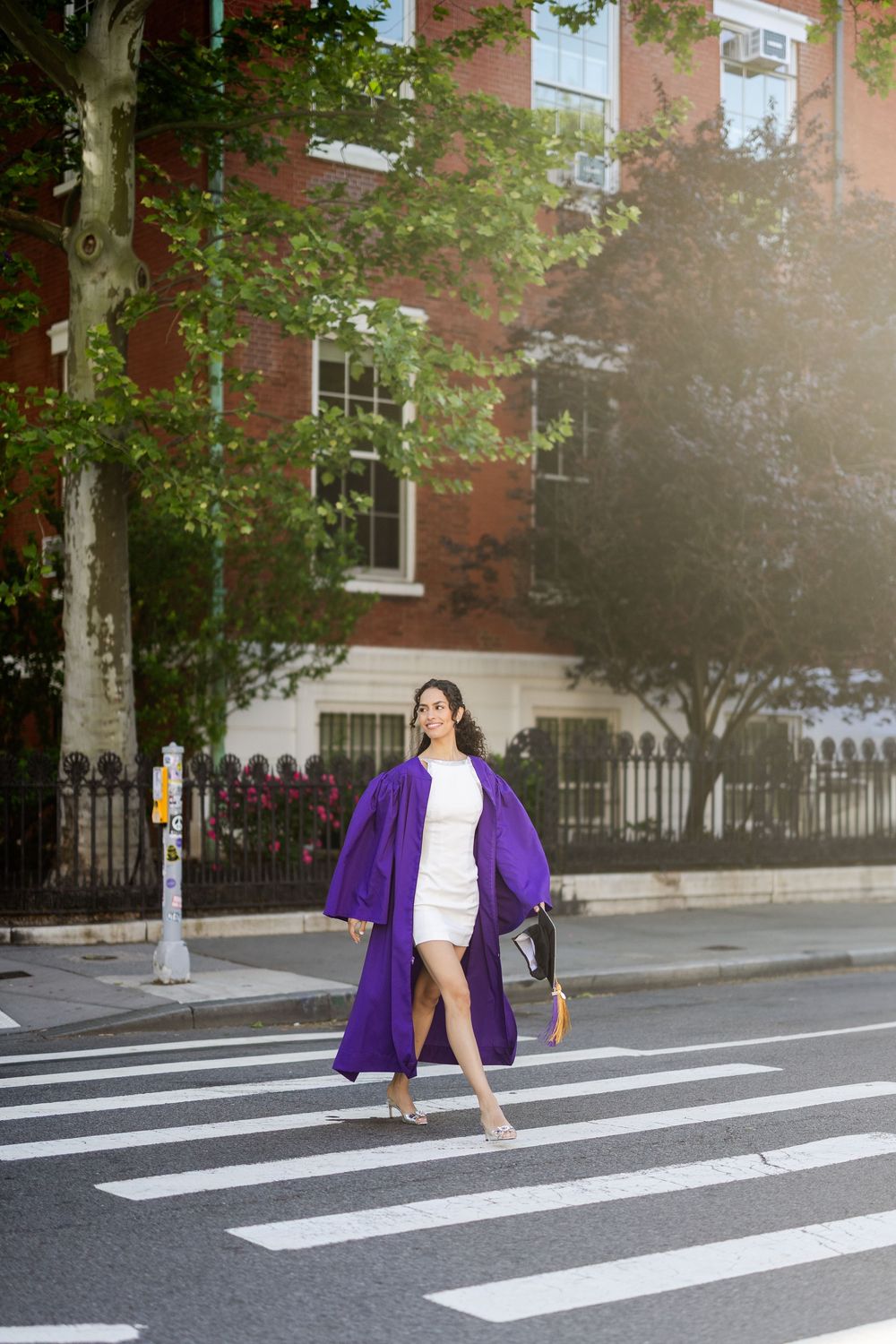 A person in a purple cardigan and white top walks across a crosswalk on a sunny urban street.