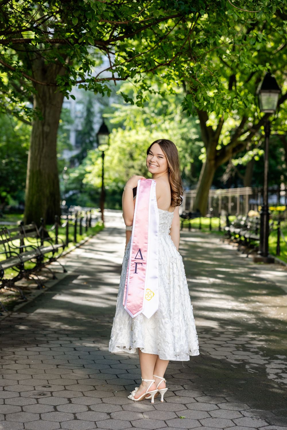A person in a white lace dress and pink sash poses along a tree-lined walkway with brick pavers.