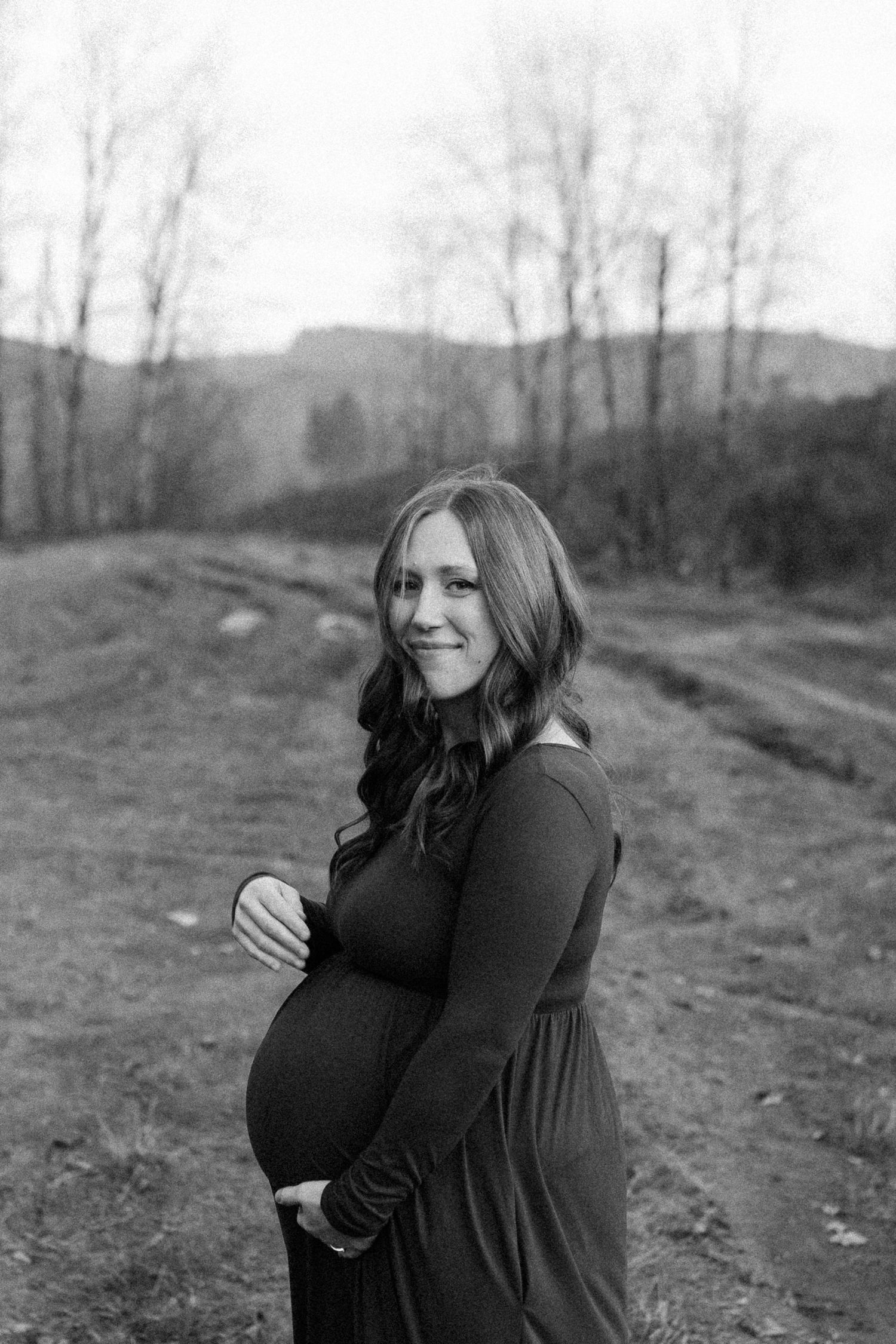 Black and white maternity portrait in a serene outdoor setting with bare trees.