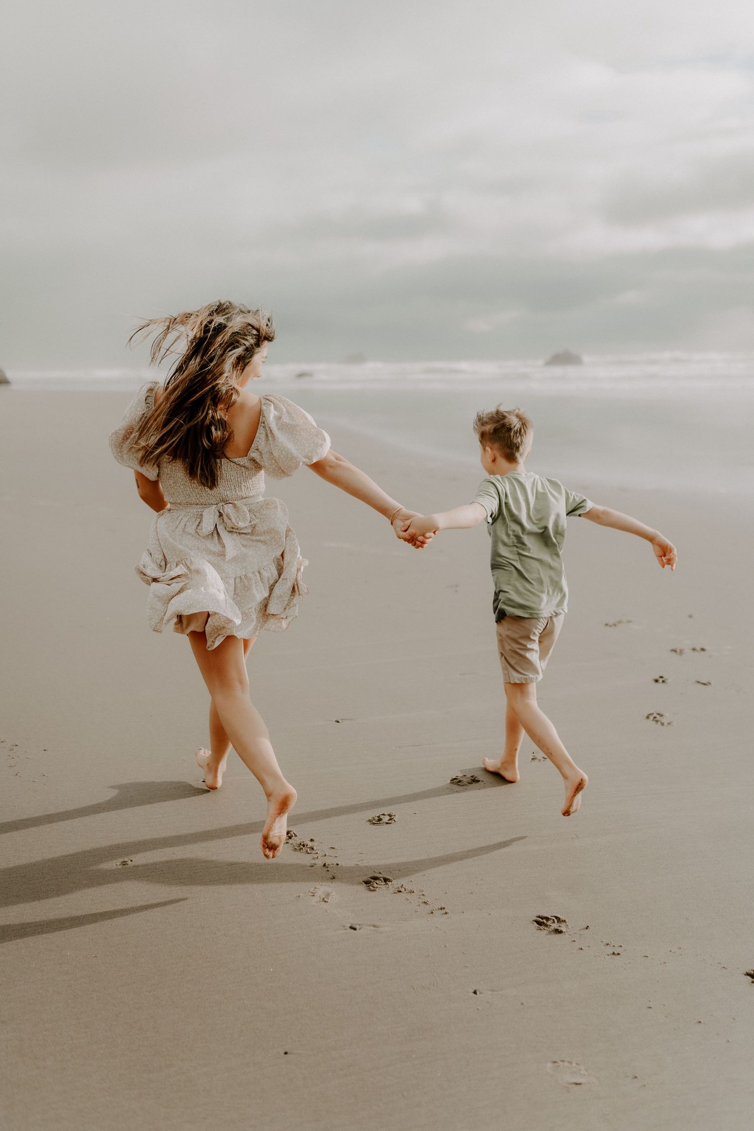 Two figures joyfully running barefoot on a misty beach at sunset, holding hands and creating footprints in the sand.