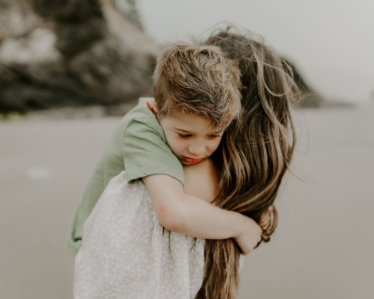 Sweet embrace between two people in casual attire on a cloudy beach with rocky cliffs in the distance.
