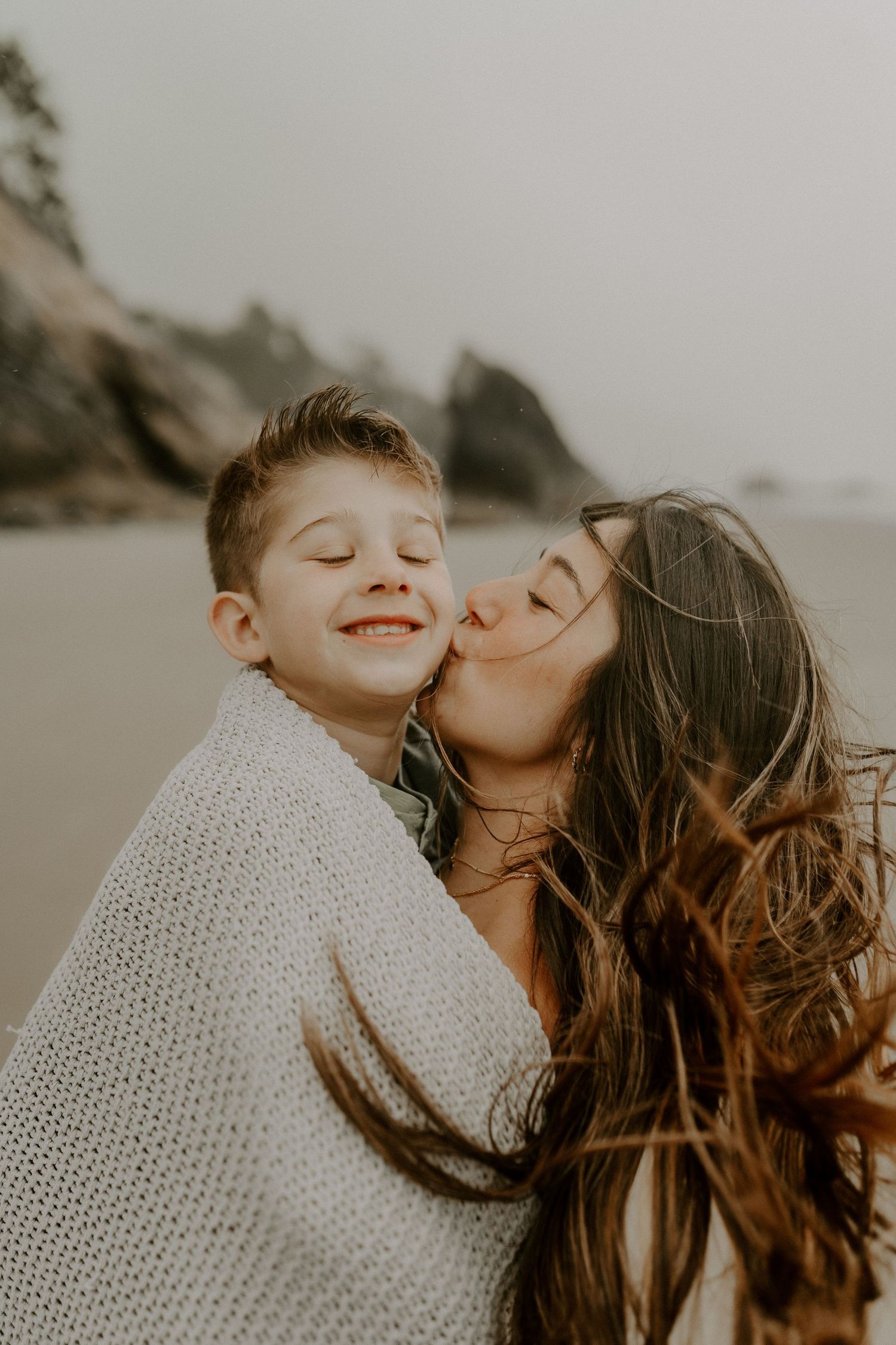 Tender moment shared between two people wrapped in a knit blanket on a cloudy beach.