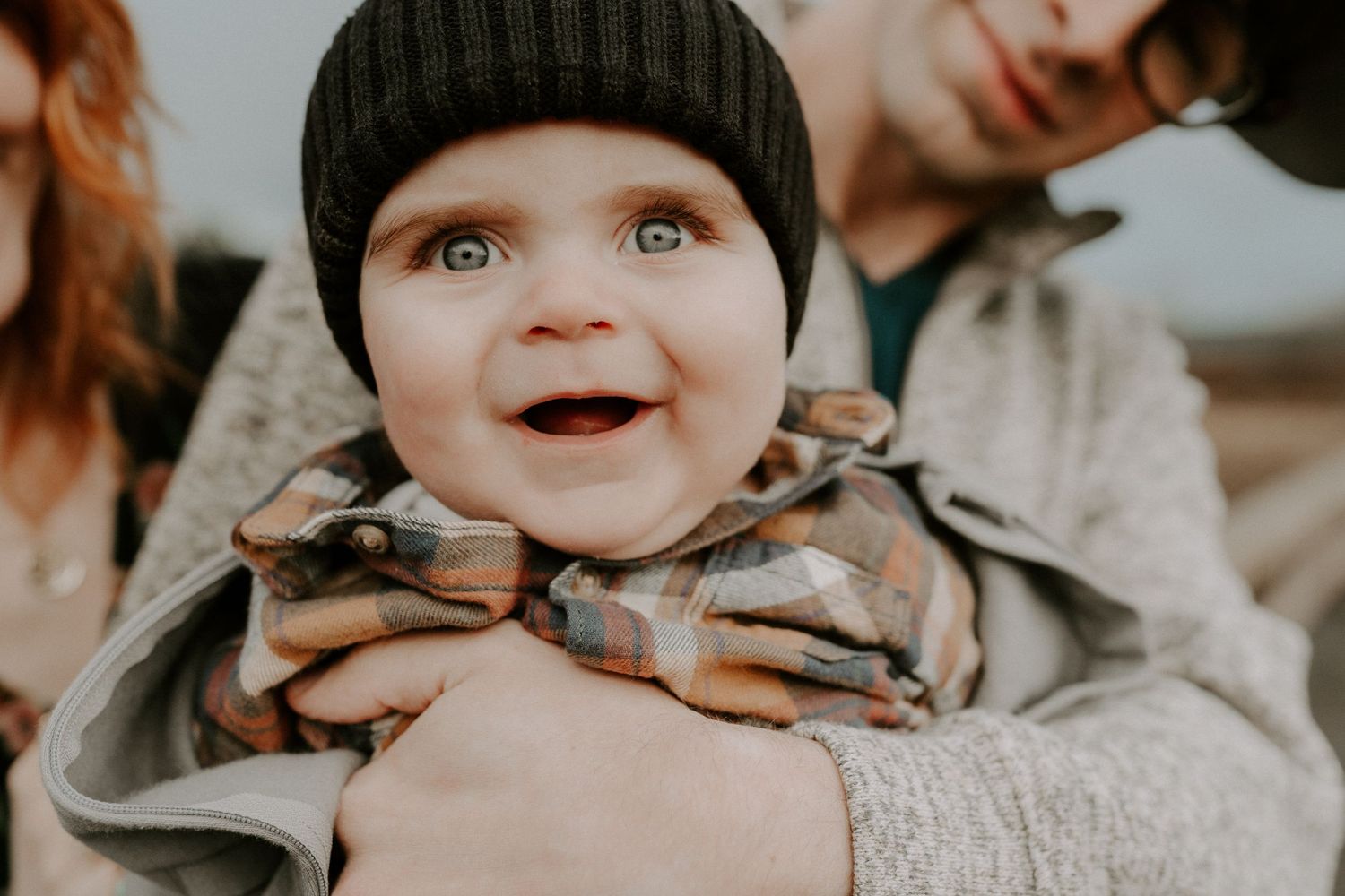 Baby wearing winter hat smiles brightly at camera while being held in cozy winter attire.