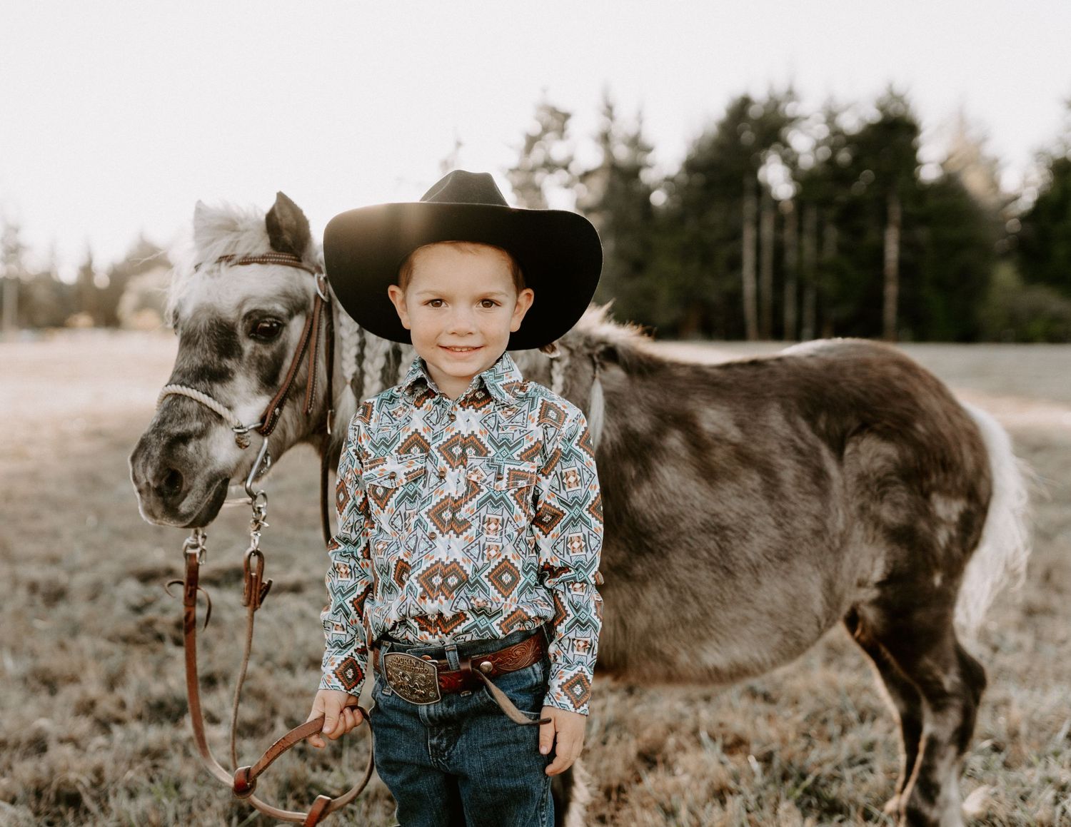 A dappled grey horse stands beside someone wearing a cowboy hat and western shirt in a rustic outdoor setting.