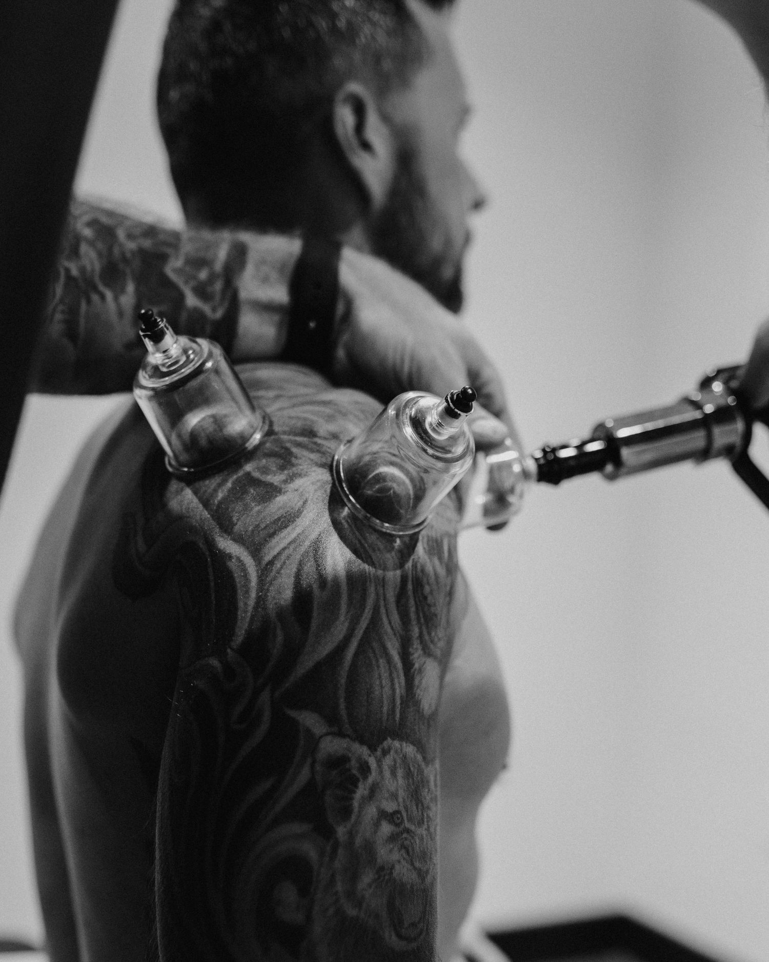 Black and white photo showing detailed tattoo work being done on someone's back.