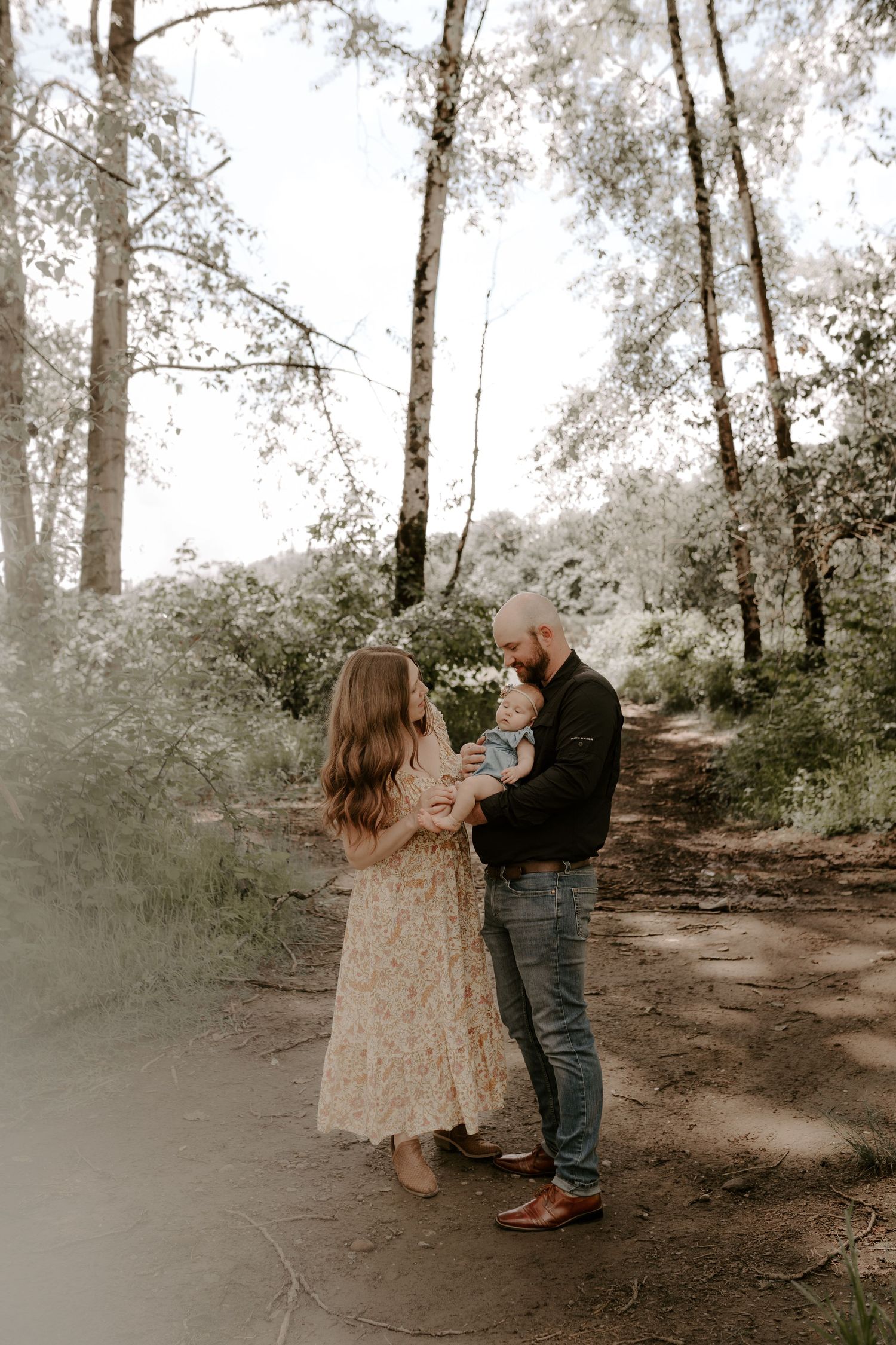 Couple standing close together on a wooded forest path with sunlight filtering through the trees.