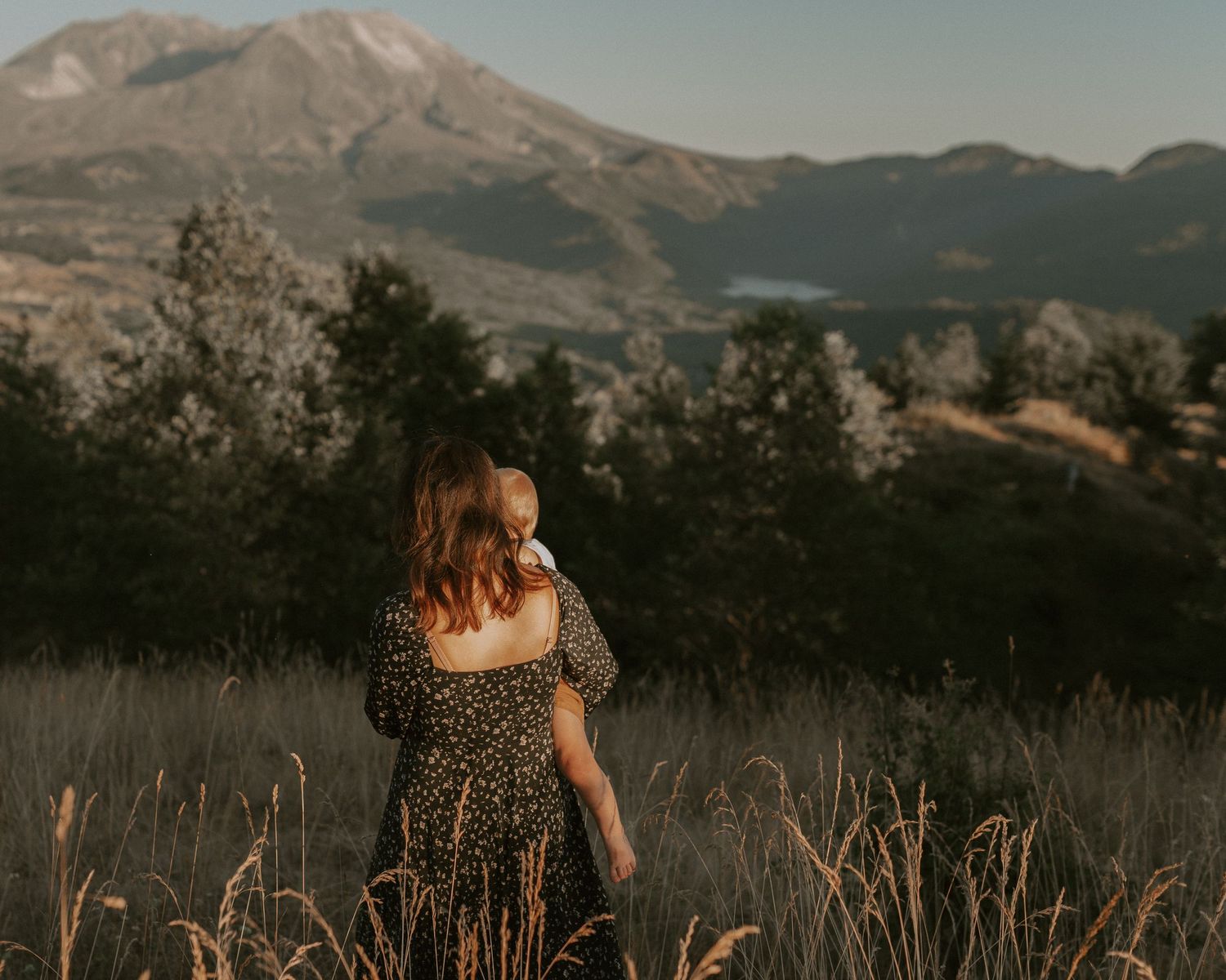 Woman in a flowing dress stands in tall grass with mountain landscape in background at golden hour.