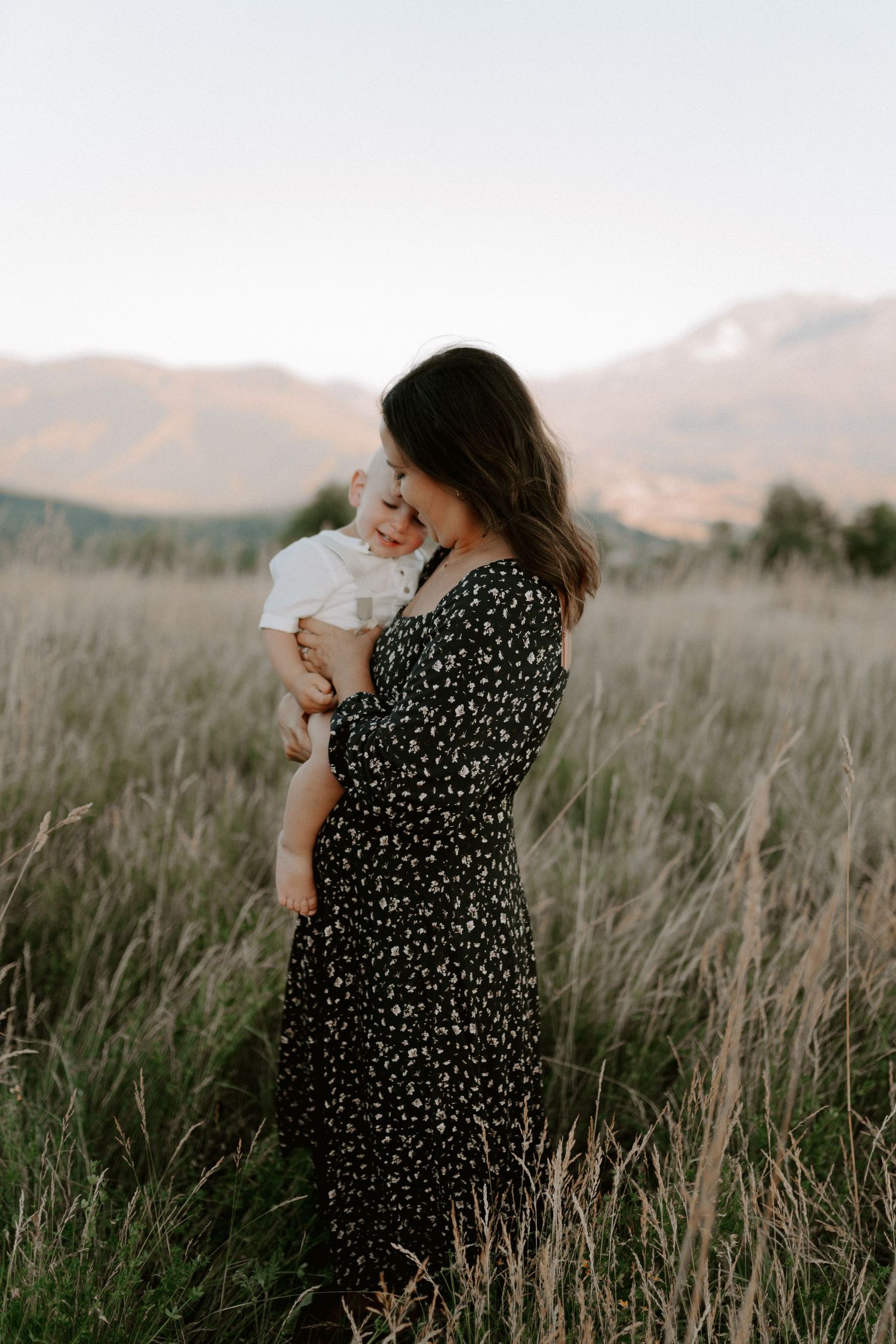 A floral dress-clad figure holds a baby in tall grass with mountains in the background at sunset.
