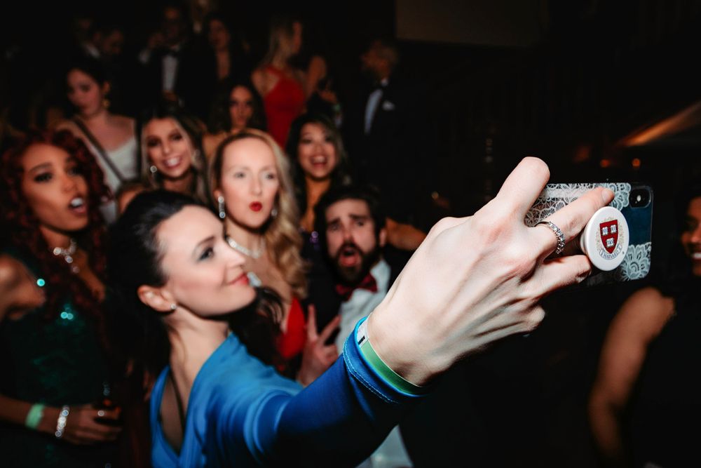 Woman in blue dress takes a selfie with a crowd of elegantly dressed guests at a formal party.