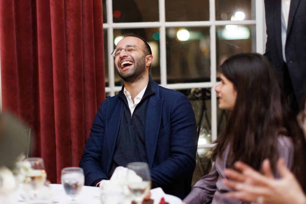 A man in a navy blazer laughs joyfully while seated at a formal dinner table with guests.