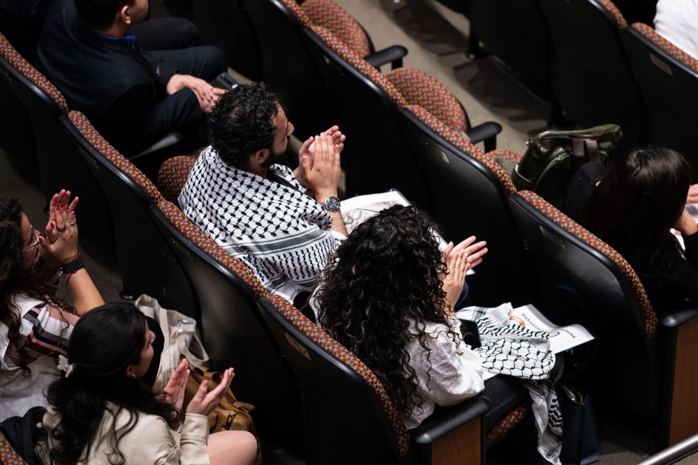 Audience members applauding at a formal event, some wearing traditional keffiyeh scarves.